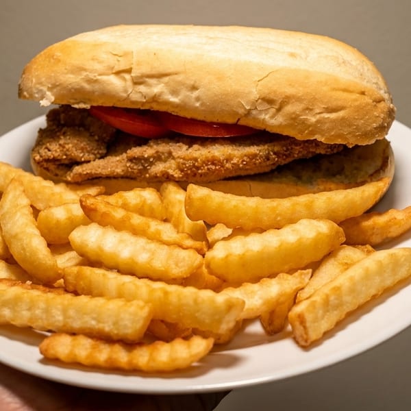 A fried fish sandwich with tomatoes on a bun, served with a side of crinkle-cut fries.