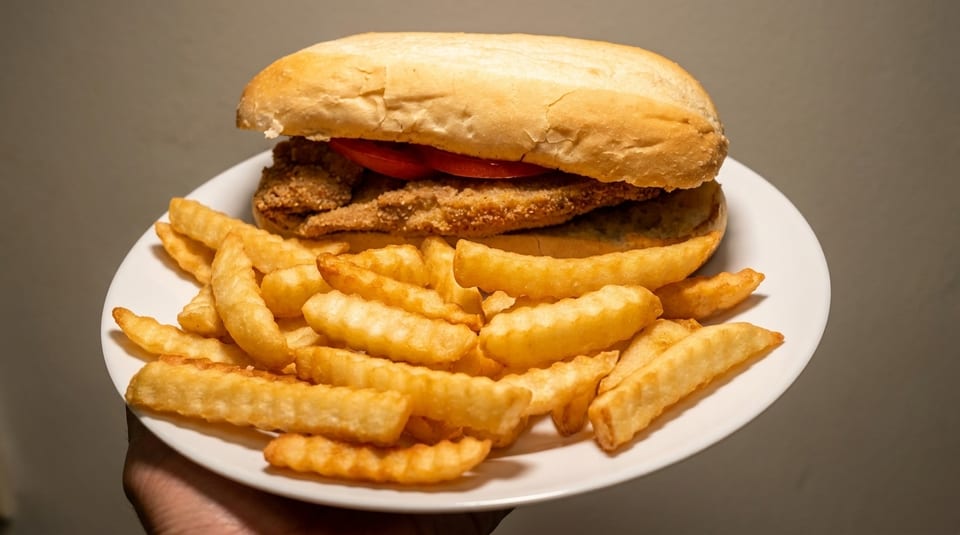 A fried fish sandwich with tomatoes on a bun, served with a side of crinkle-cut fries.