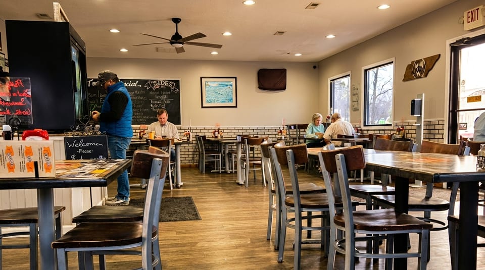 A casual restaurant interior with customers seated at tables and a person standing near a chalkboard menu.
