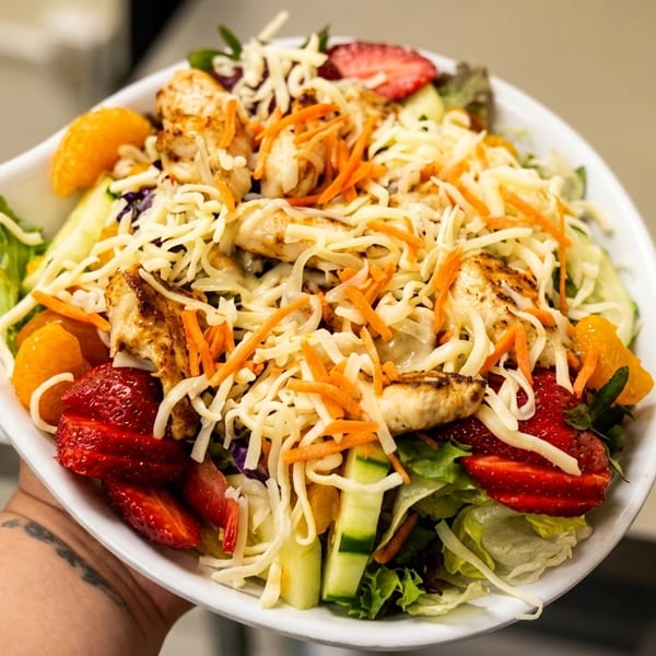 A vibrant salad with grilled chicken, strawberries, mandarin oranges, shredded cheese, and carrots.